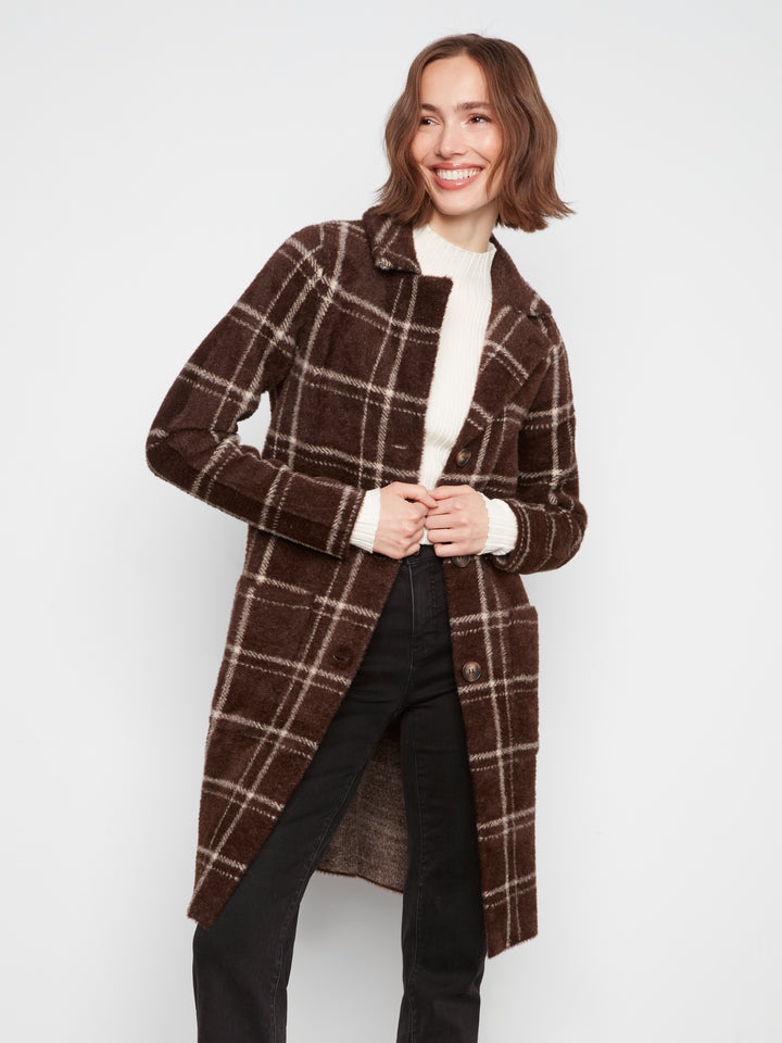A woman modeling a brown and beige plaid mid-length coat over a cream turtleneck and black pants, photographed against a plain light gray studio background.