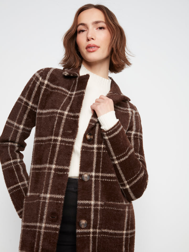 A woman wearing a brown wool-blend plaid coat over a white turtleneck sweater and black trousers, photographed against a plain light gray studio background.