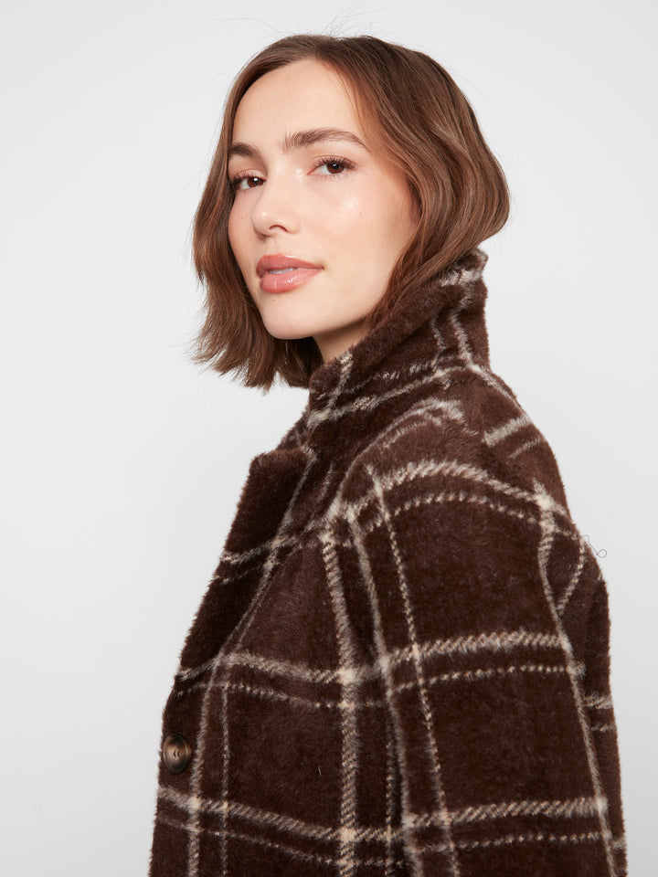 A woman with a short brown bob haircut is shown in profile wearing a brown fuzzy plaid coat with cream lines, set against a plain light gray studio background.