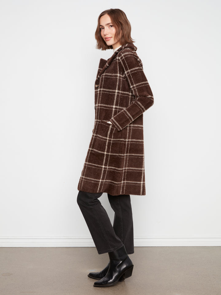 A woman modeling a brown plaid coat with black trousers and black ankle boots, standing against a plain white wall.