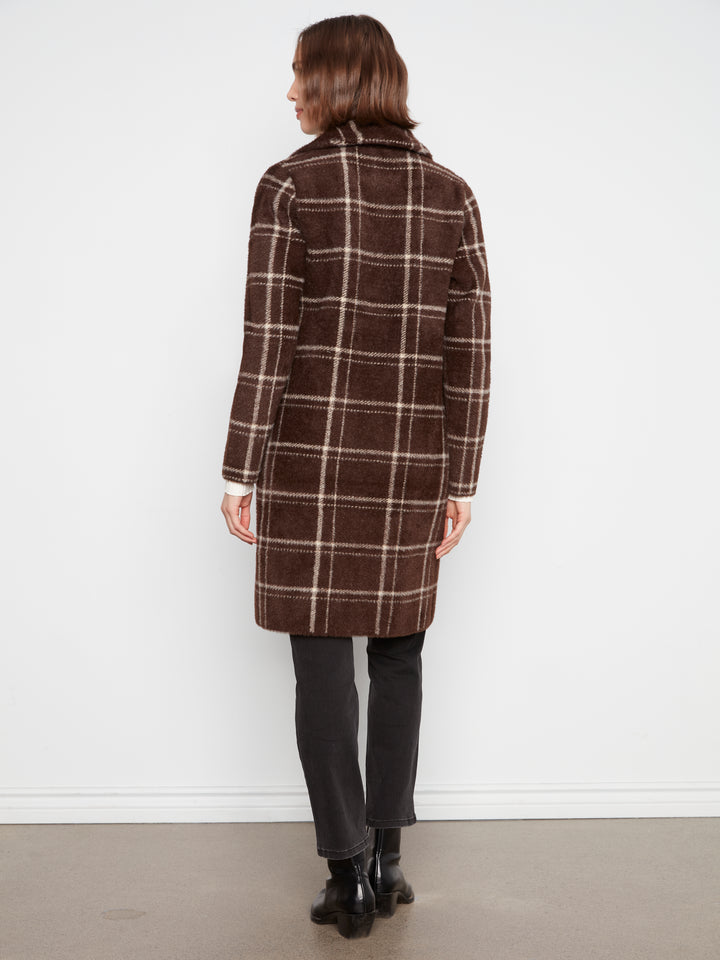 A woman viewed from the back wearing a brown plaid coat, dark trousers, and black ankle boots, standing against a plain white wall.