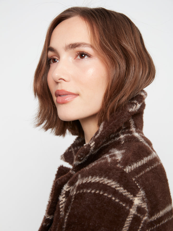 A woman with a short brown bob haircut wearing a brown fuzzy knit sweater with a cream plaid pattern, shown in a close-up profile against a light gray background.