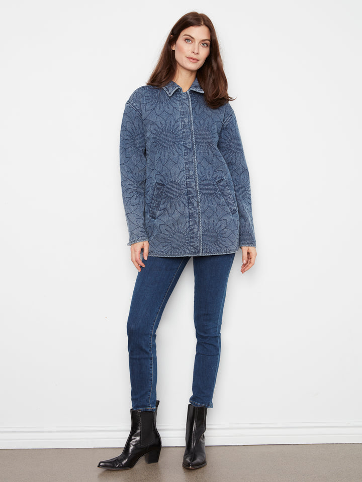 A woman modeling a blue denim jacket with an all-over tonal floral jacquard pattern, paired with dark blue skinny jeans and black ankle boots, standing against a plain white wall.