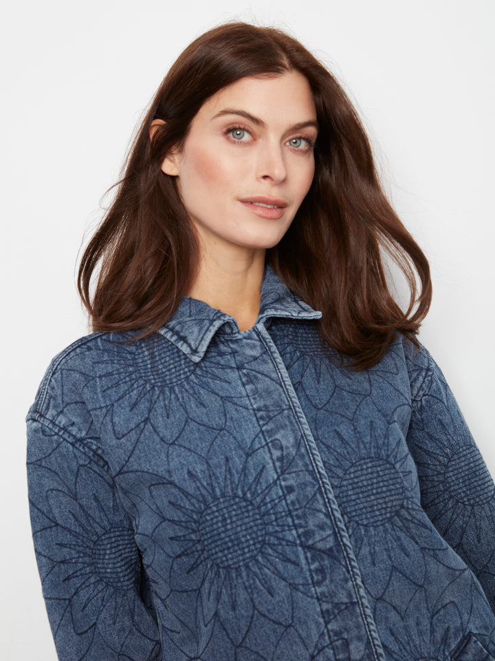 A woman with long brown hair wearing a textured blue denim jacket with a sunflower-like embossed pattern, photographed against a plain light gray background.
