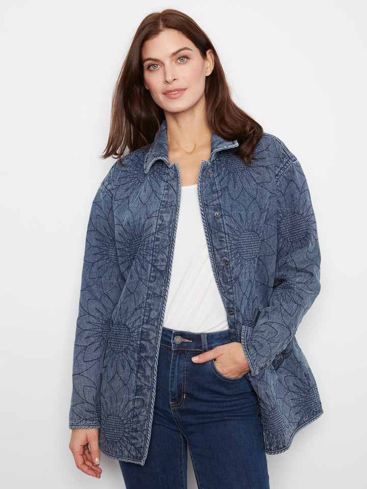 A woman wearing a blue patterned button-up shirt jacket over a white top and dark blue jeans, photographed against a plain light background.