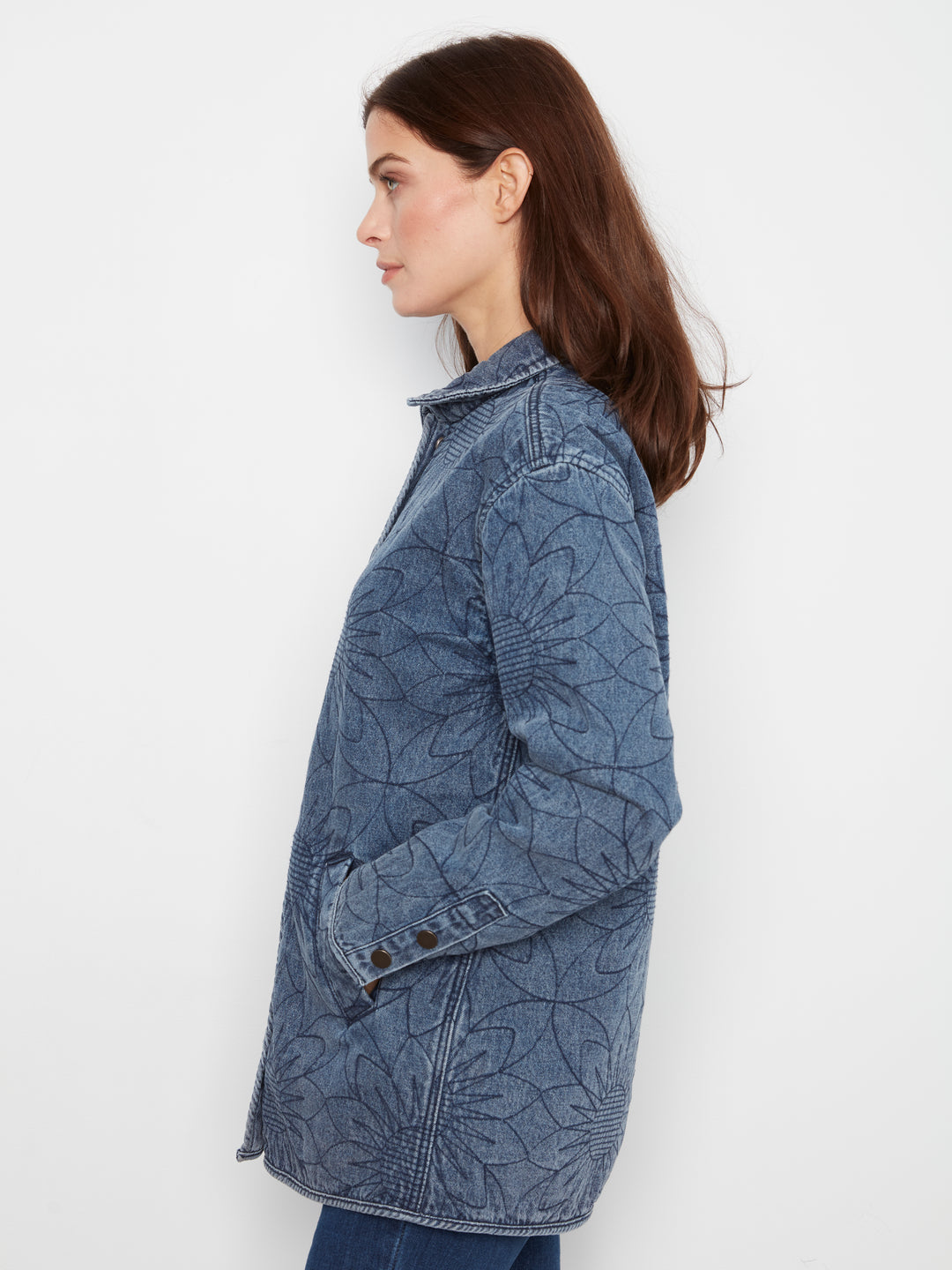 A woman shown in side profile wearing a blue denim jacket with an allover tonal floral or leaf-like embossed pattern, against a plain white studio background.