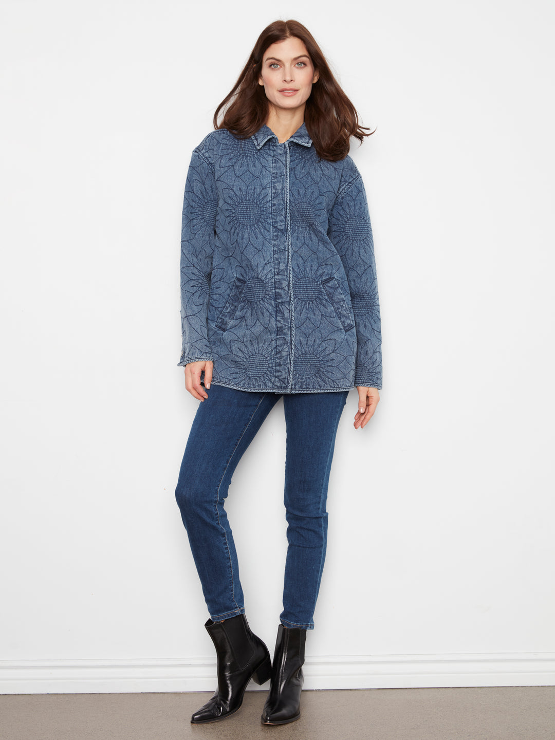 A woman modeling a blue textured jacket with a mandala-like pattern, paired with dark blue skinny jeans and black ankle boots, standing against a plain white wall.