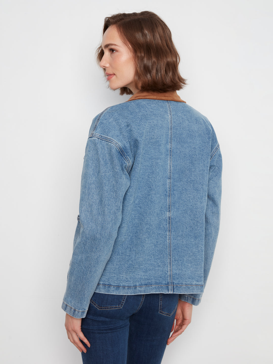 A woman shown from the back wearing a light blue denim jacket with a brown collar, paired with dark blue jeans, against a plain white studio background.