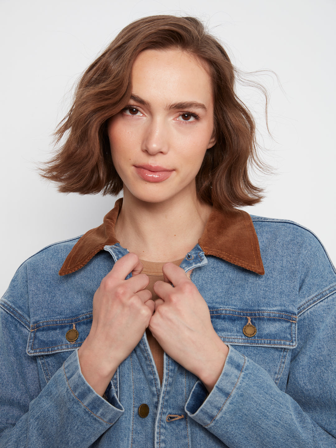 A woman with a short brown wig wearing a blue denim jacket with a brown suede collar, posing against a light gray background.