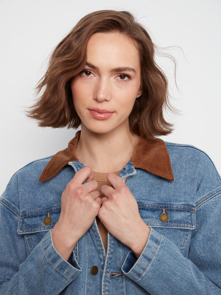A woman with a short brown wig wearing a blue denim jacket with a brown suede collar, posing against a light gray background.