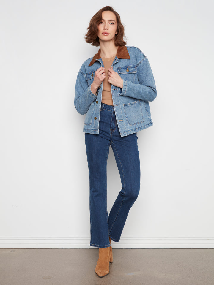 A woman modeling a light blue denim jacket with a brown corduroy collar, paired with dark blue jeans and tan ankle boots, standing against a white wall.