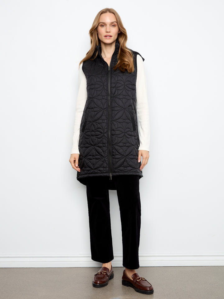 A woman stands against a plain white wall wearing a black quilted zip-front vest over a white long-sleeve top, black pants, and brown loafers.