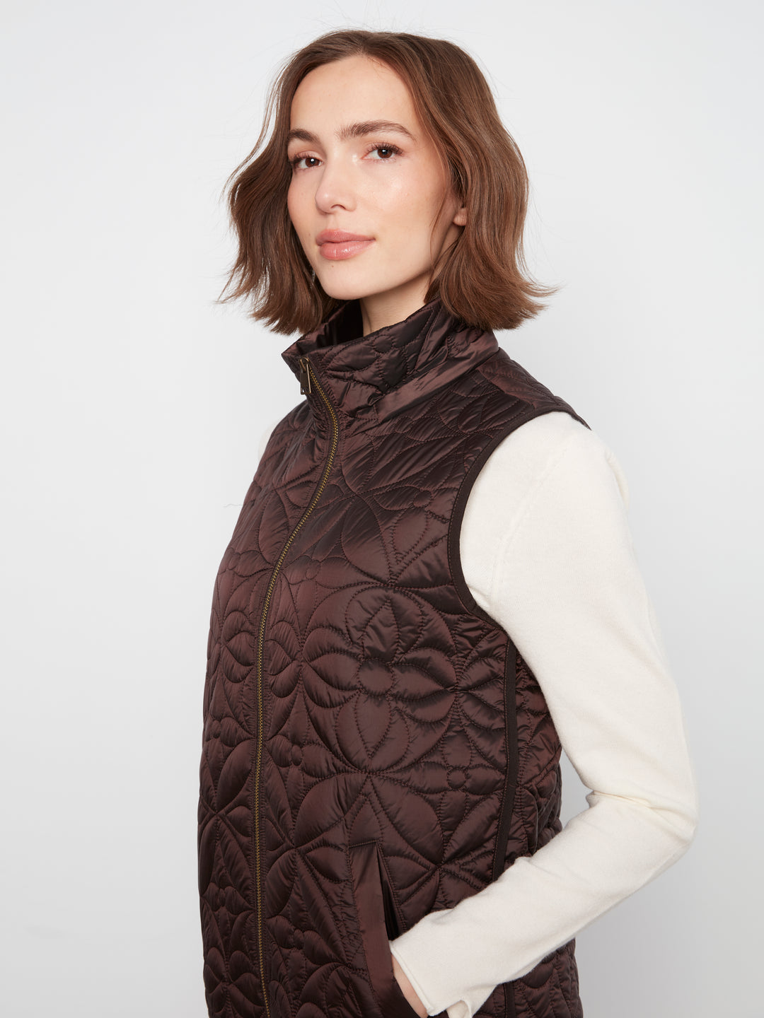 A woman wearing a dark brown quilted puffer vest over a cream long-sleeve top, photographed against a plain light gray studio background.