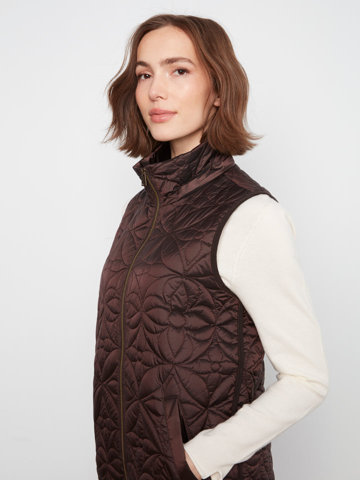 A woman wearing a dark brown quilted puffer vest over a cream long-sleeve top, photographed against a plain light gray studio background.