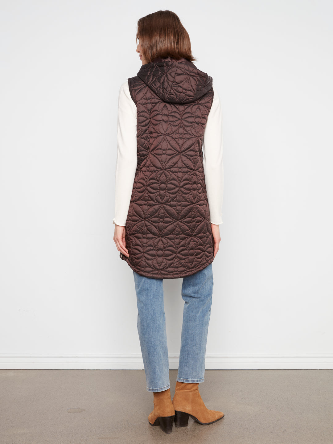 A woman shown from the back wearing a dark brown quilted hooded puffer vest over a white long-sleeve top, blue jeans, and tan ankle boots, standing against a plain white wall.
