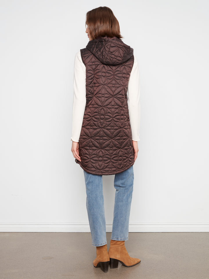 A woman shown from the back wearing a dark brown quilted hooded puffer vest over a white long-sleeve top, blue jeans, and tan ankle boots, standing against a plain white wall.
