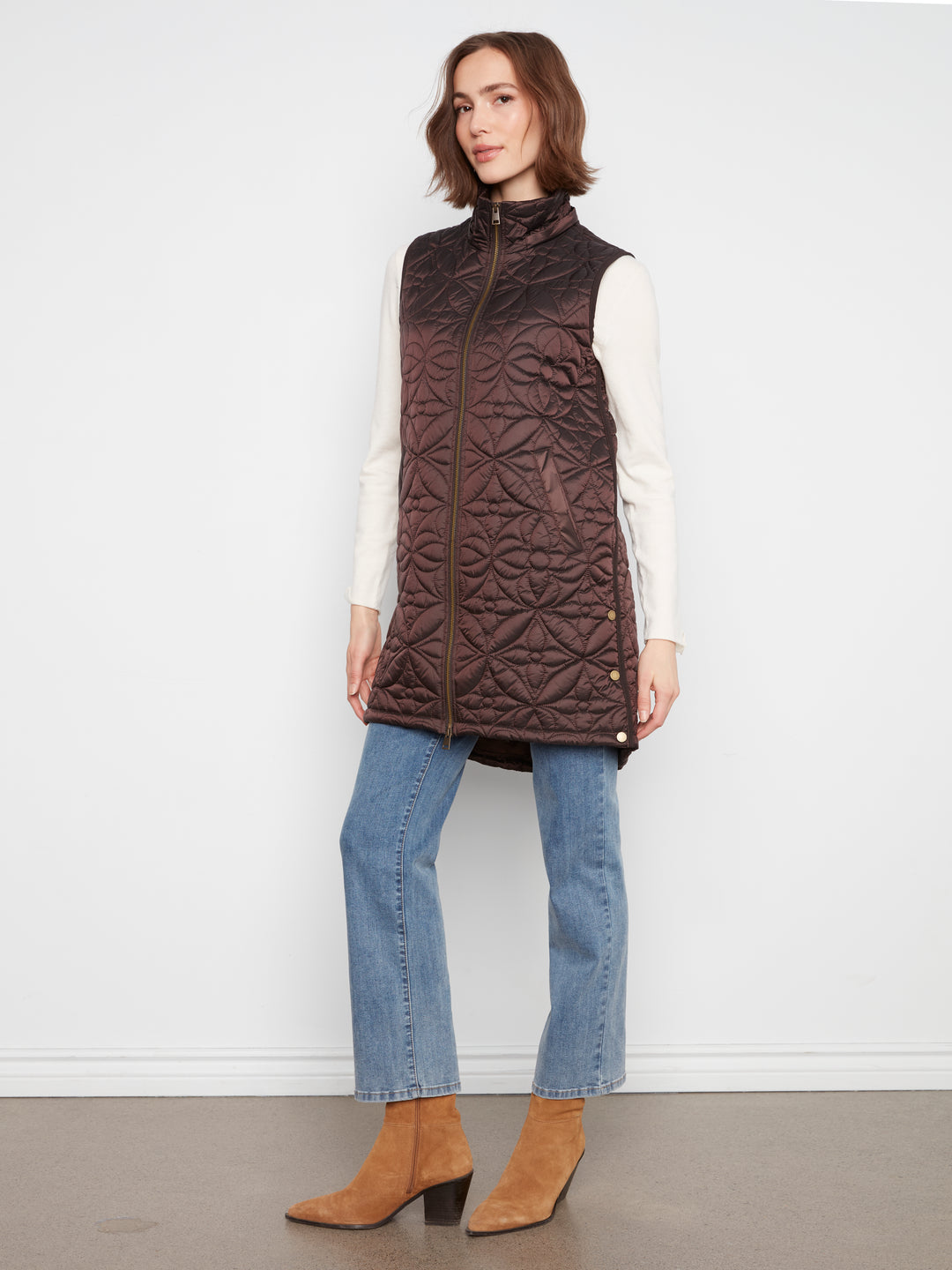 A woman models a dark brown quilted sleeveless puffer vest over a white long-sleeve top, paired with light blue jeans and tan ankle boots, standing against a plain white wall.