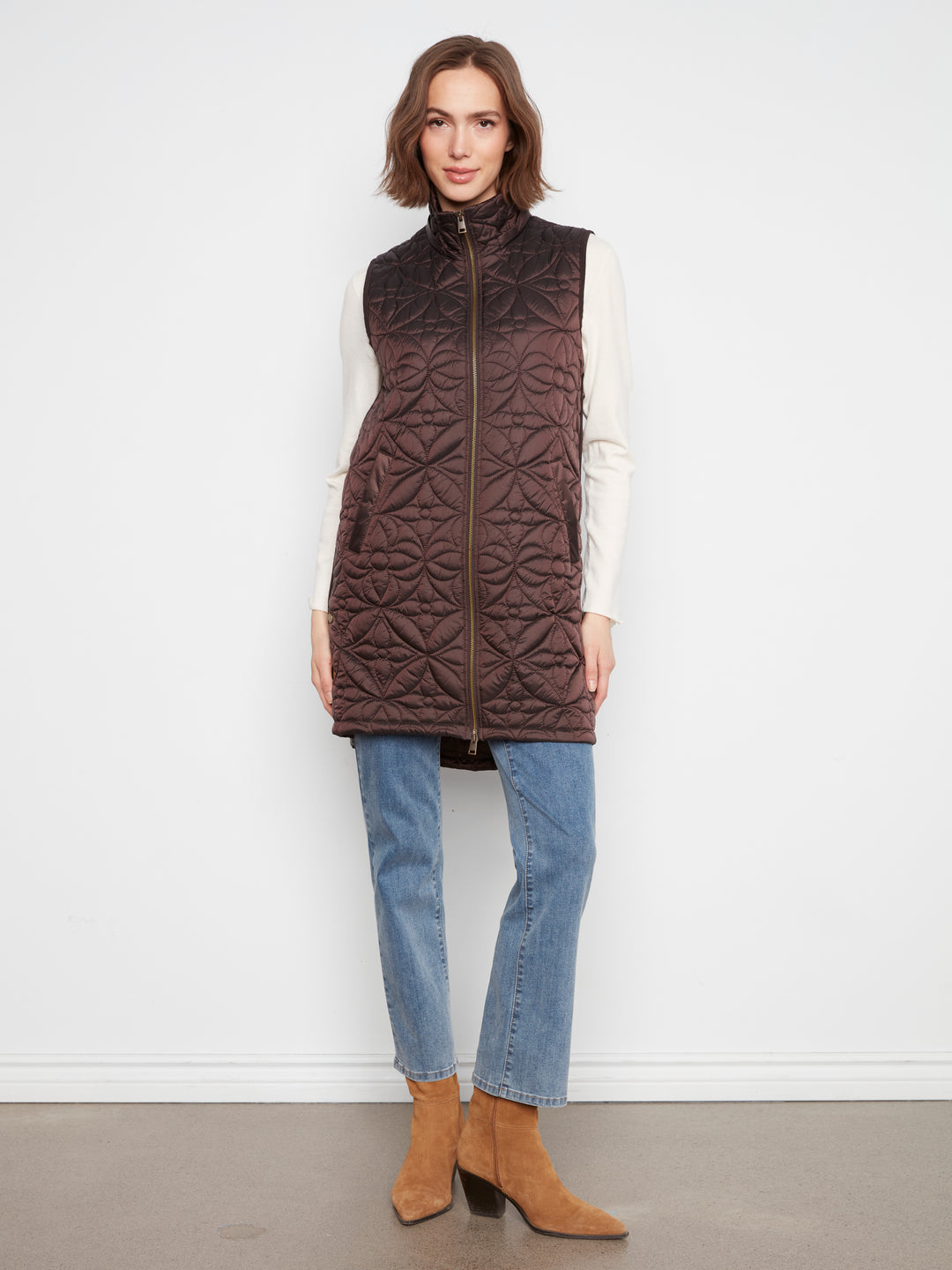 A woman modeling a dark brown quilted zip-front puffer vest over a white long-sleeve top, blue jeans, and tan ankle boots, standing against a plain white wall.