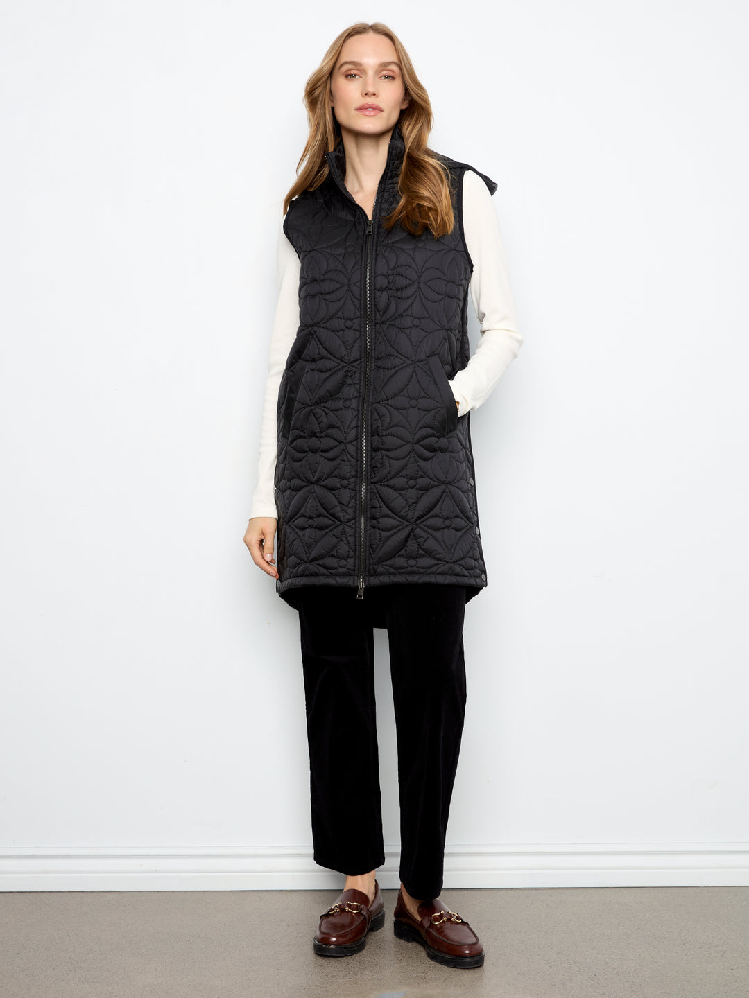 A woman stands against a plain white wall wearing a black quilted zip-front vest over a white long-sleeve top, black pants, and brown loafers.