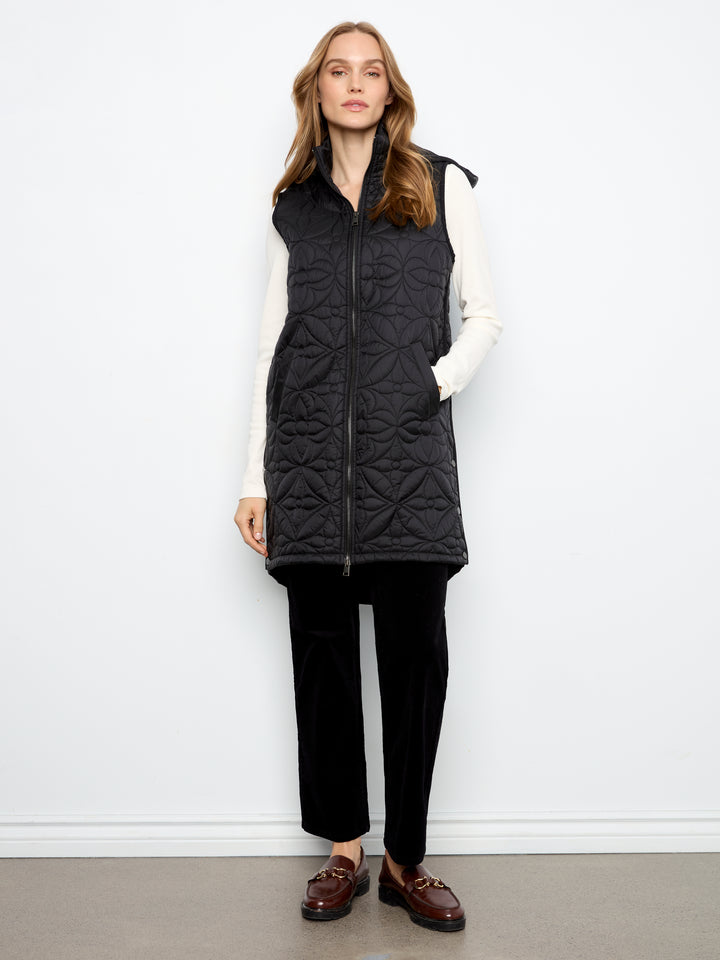 A woman stands against a plain white wall wearing a black quilted zip-front vest over a white long-sleeve top, black pants, and brown loafers.