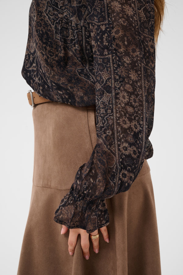A close-up of a person wearing a sheer black and brown paisley patterned blouse with long ruffled sleeves, paired with a brown skirt.