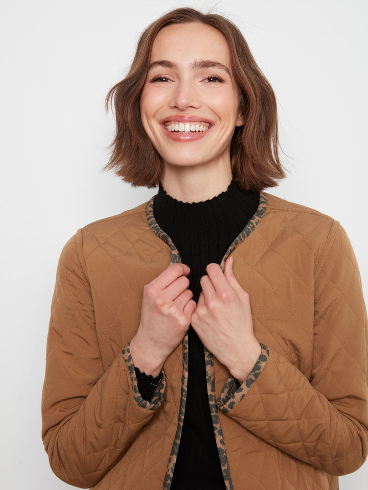 A smiling woman with short brown hair wearing a tan quilted jacket with patterned trim over a black top, photographed against a plain light gray background.