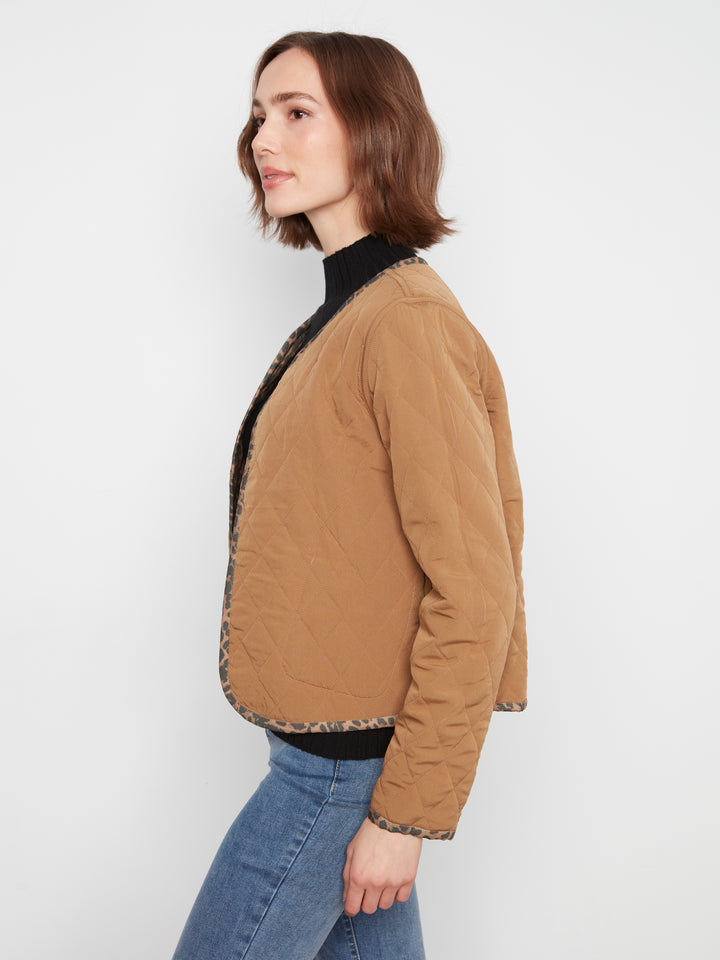 A woman shown in side profile wearing a tan quilted jacket with a black turtleneck underneath and blue jeans, photographed against a plain light gray studio background.