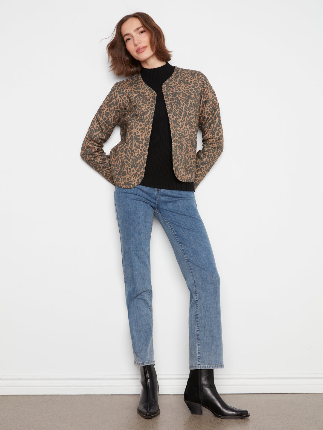 A woman modeling a cropped leopard-print jacket over a black top, paired with light-wash blue jeans and black ankle boots, standing against a plain white wall.