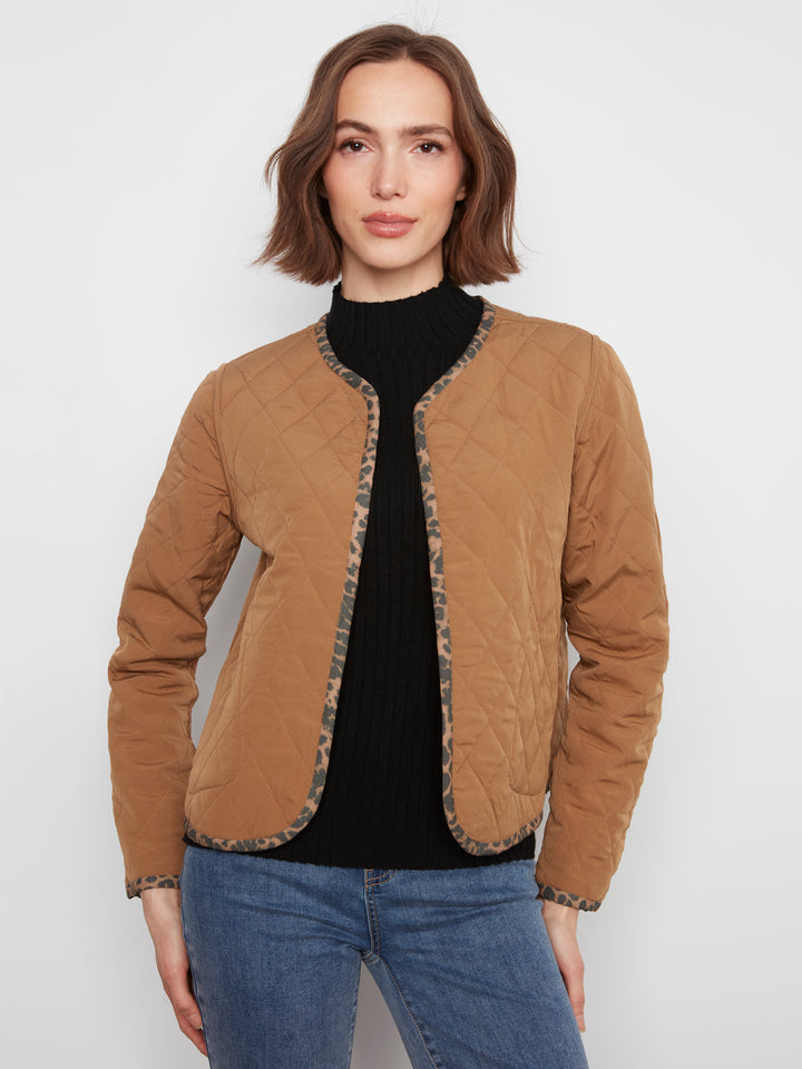 A woman wearing a tan quilted jacket with patterned trim over a black turtleneck and blue jeans, standing against a light gray studio background.
