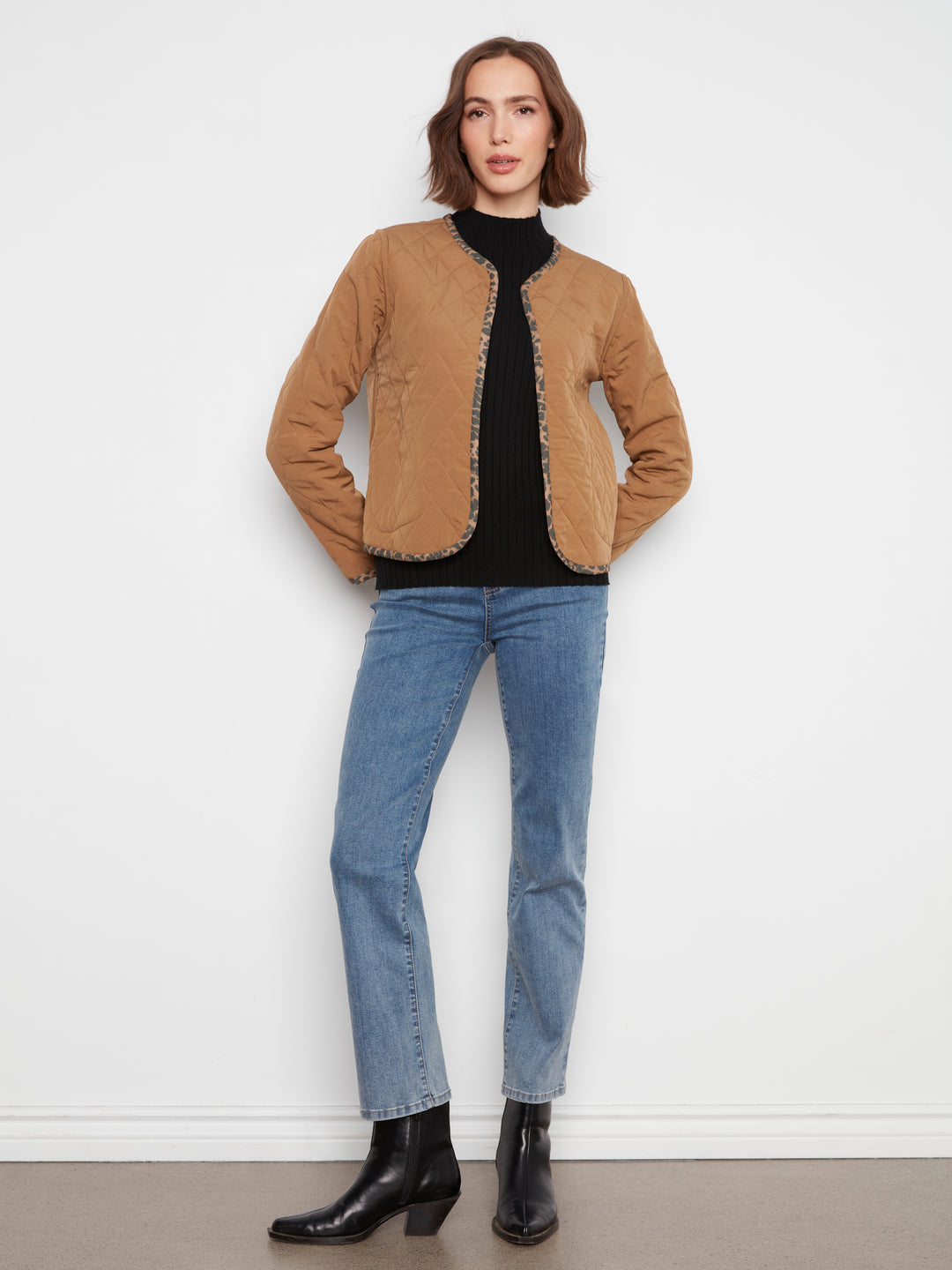 A woman stands against a plain white wall wearing a tan suede jacket with decorative trim, a black turtleneck, light blue jeans, and black ankle boots.