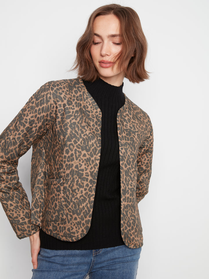 A woman wearing a brown and beige leopard-print blazer over a black ribbed turtleneck sweater and blue jeans, photographed against a plain light gray studio background.