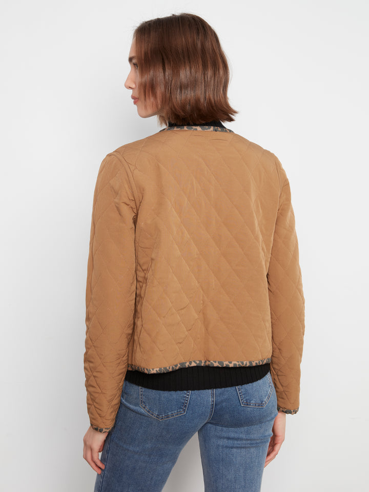 A woman shown from the back wearing a tan quilted jacket with patterned trim and blue jeans, photographed against a plain light gray studio background.