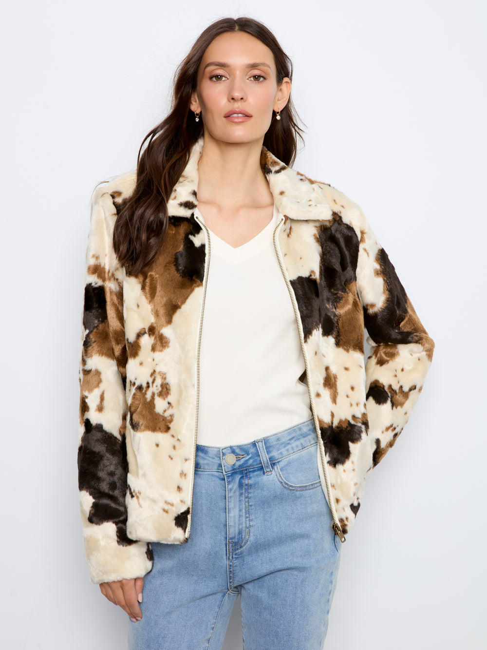 A woman modeling a faux fur jacket with a brown, black, and cream cow print pattern, worn over a white top and blue jeans against a light gray background.
