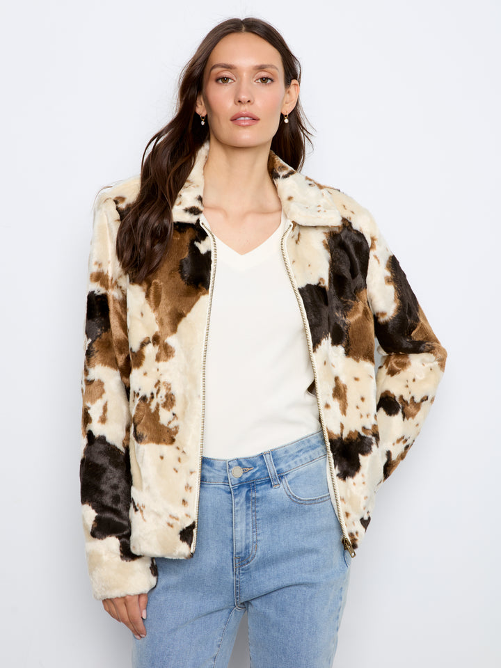 A woman modeling a faux fur jacket with a brown, black, and cream cow print pattern, worn over a white top and blue jeans against a light gray background.