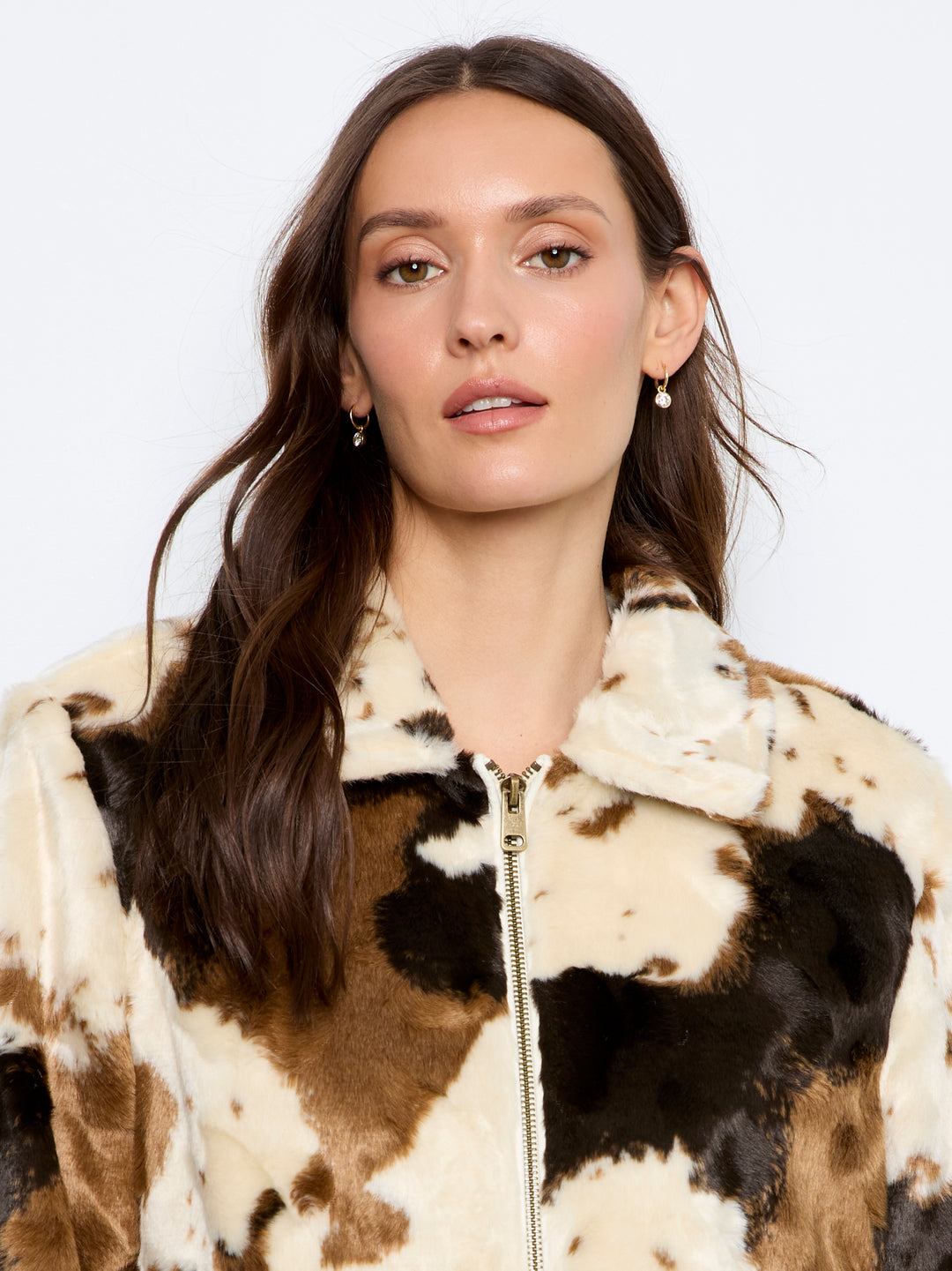 A woman wearing a brown, black, and cream cow-print faux fur jacket with a gold zipper, photographed against a plain white background.