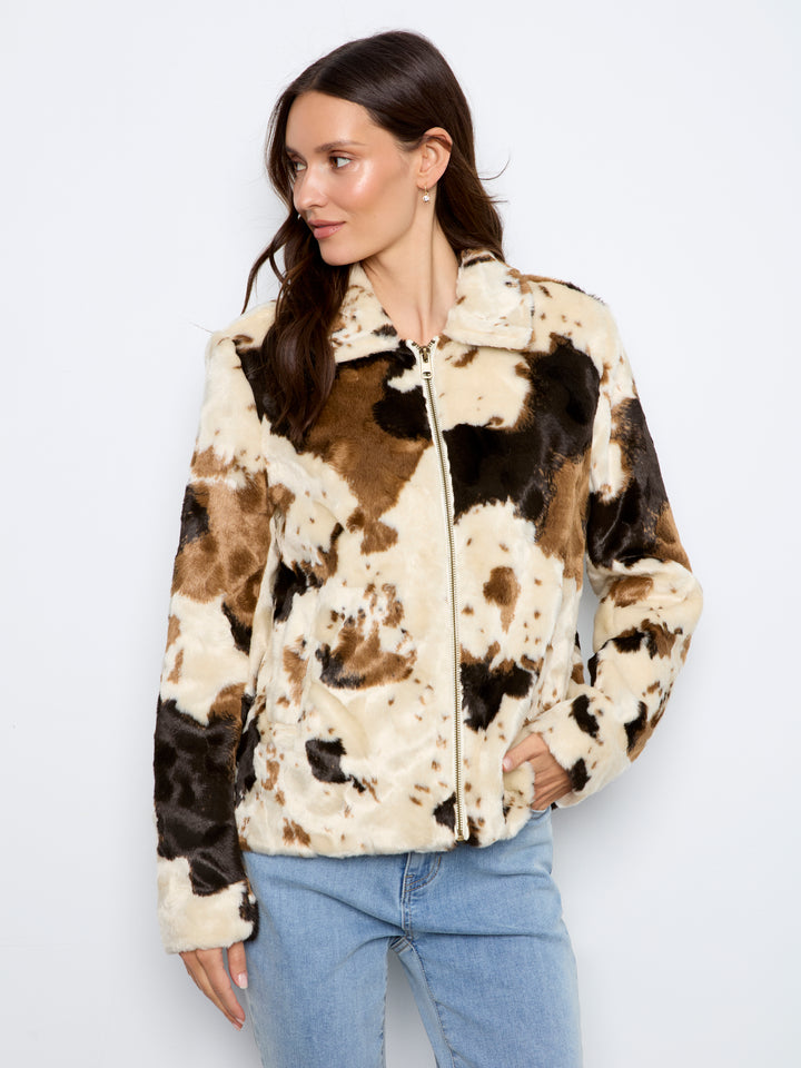 A woman wearing a faux fur zip-up jacket with a brown, black, and beige cow-print pattern, paired with light blue jeans, against a plain white background.