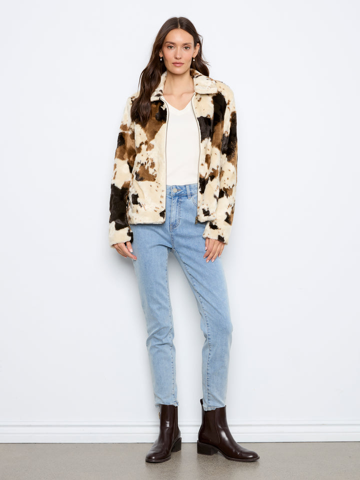 A woman modeling a cropped patchwork faux fur jacket with brown, black, and cream patches, worn over a white top and light blue jeans, standing against a white wall.