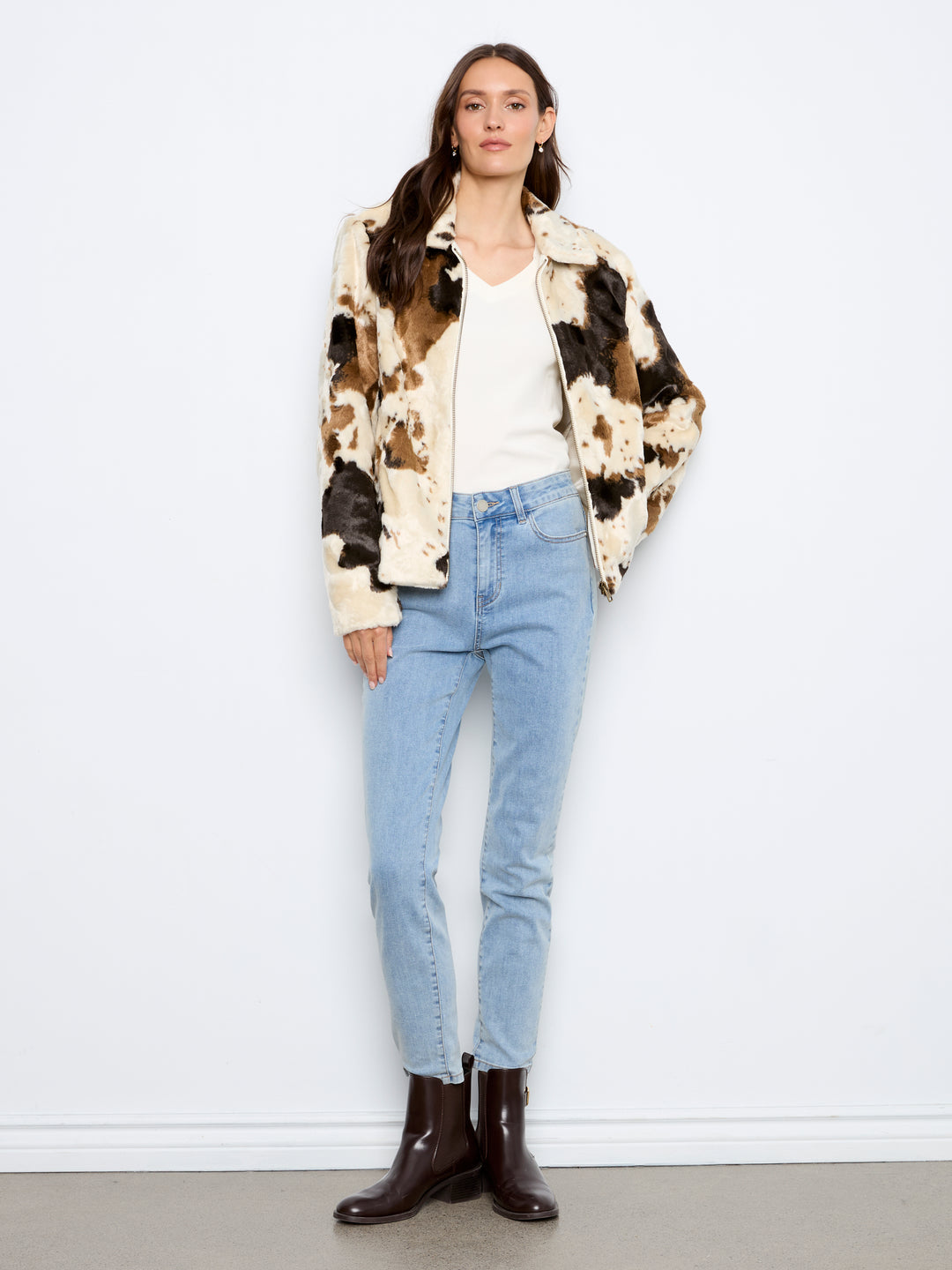 A woman stands against a plain white wall wearing a cream, brown, and black cow-print jacket over a white top, light blue jeans, and dark brown ankle boots.