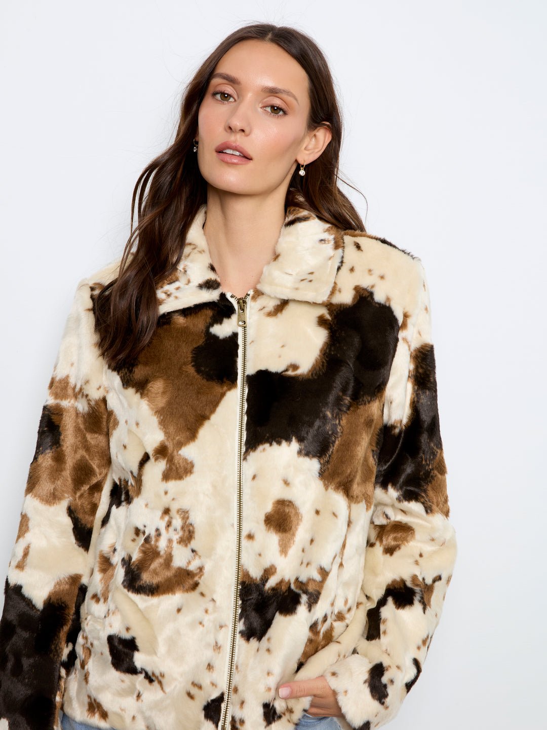 A woman wearing a faux fur zip-up jacket with a brown, beige, and black cow print pattern, photographed against a plain white studio background.