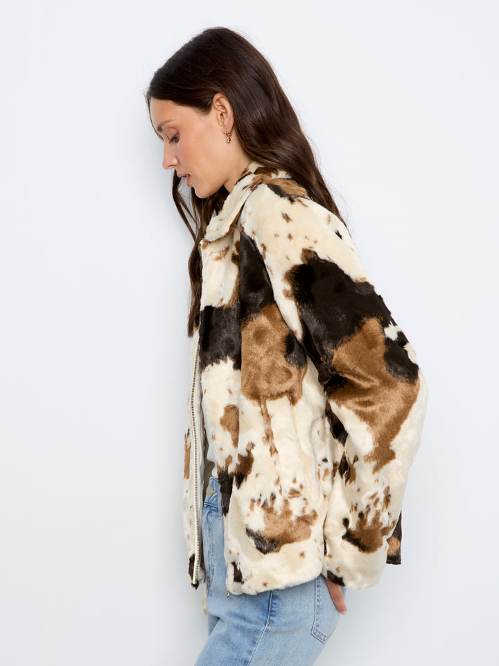 A woman wearing a cow-print faux fur jacket in brown, black, and cream, paired with light blue jeans, shown from a side profile against a plain white background.