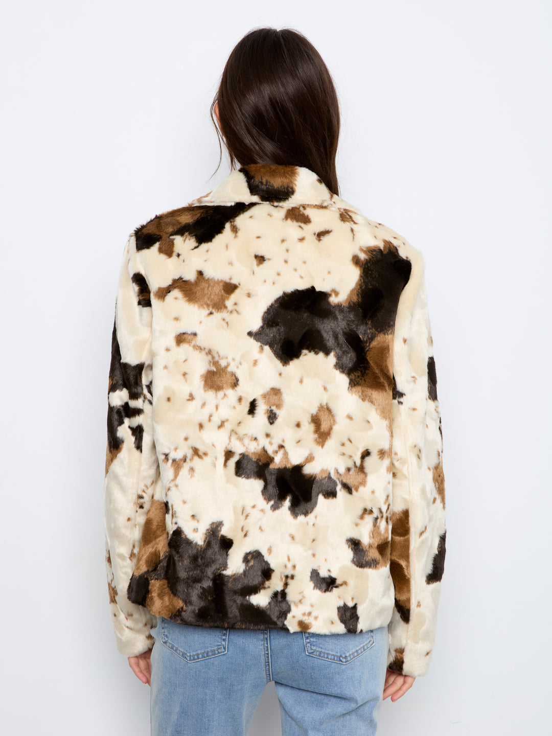 A person shown from the back wearing a faux-fur jacket with a cow-print pattern in cream, brown, and black, paired with light blue jeans against a plain white background.