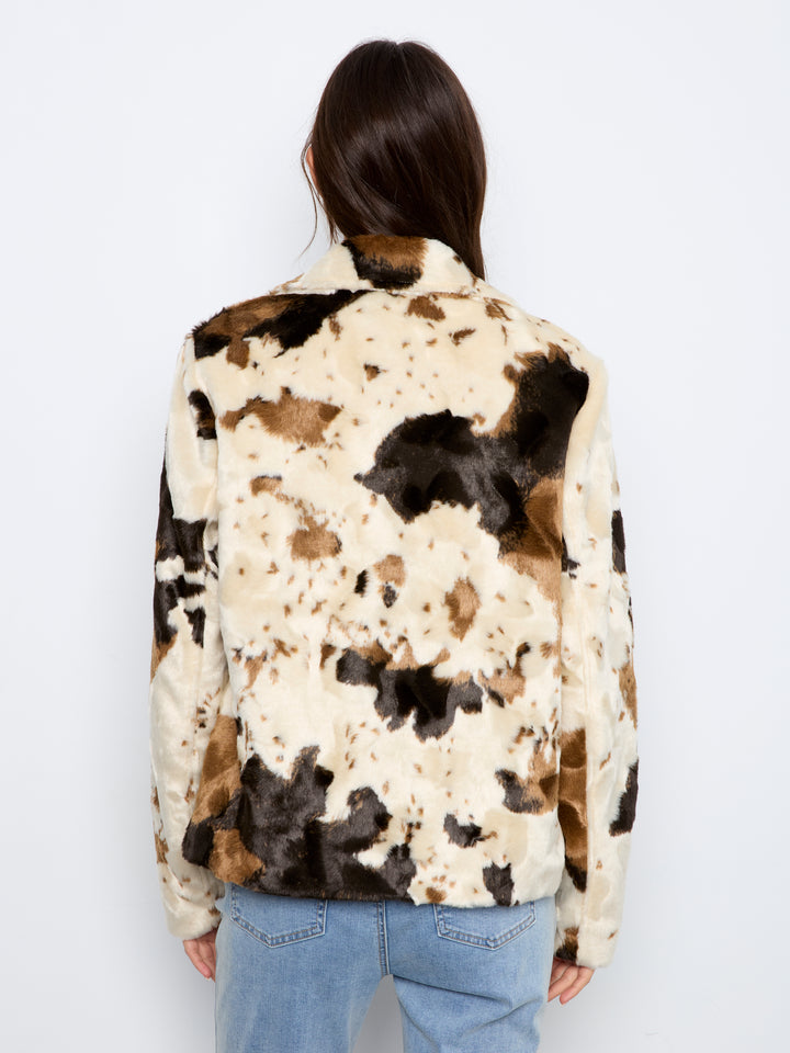 A person shown from the back wearing a faux-fur jacket with a cow-print pattern in cream, brown, and black, paired with light blue jeans against a plain white background.