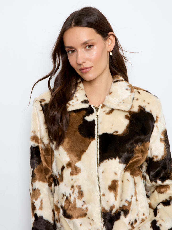 A woman wearing a faux fur zip-up jacket with a brown, black, and cream cow print pattern, photographed against a plain white studio background.