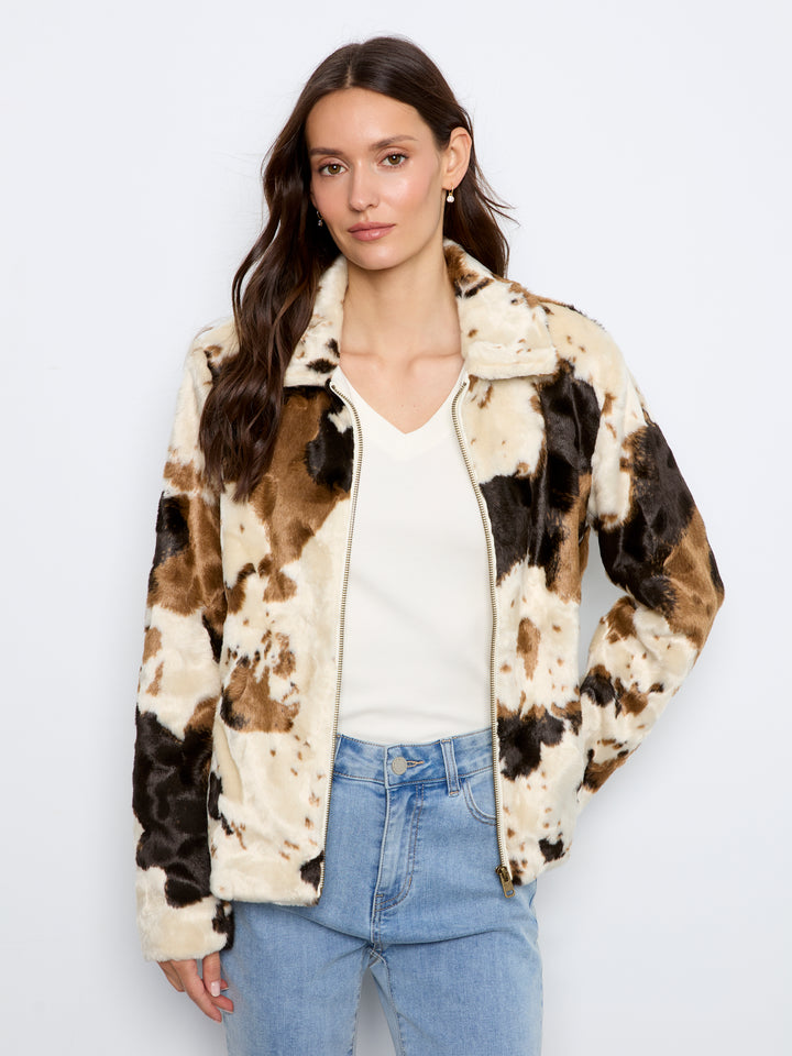 A woman modeling a fuzzy faux-fur zip-up jacket with a brown, black, and cream cow-print pattern, worn over a white top and light blue jeans against a plain white background.