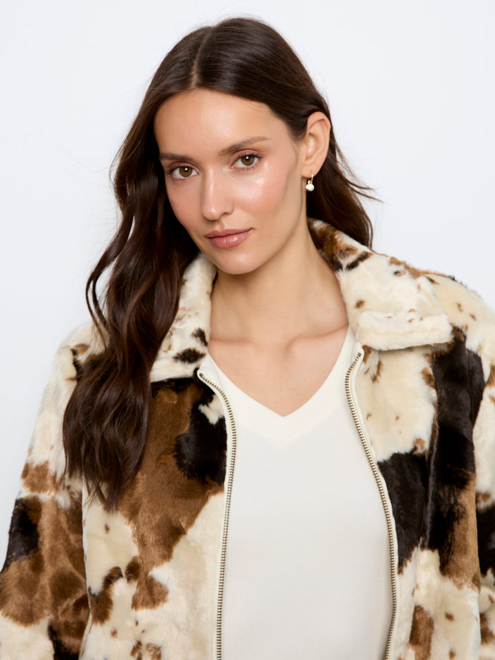 A woman wearing a faux fur zip-up jacket with a brown, black, and cream cow-print pattern over a cream V-neck top, photographed against a plain white studio background.