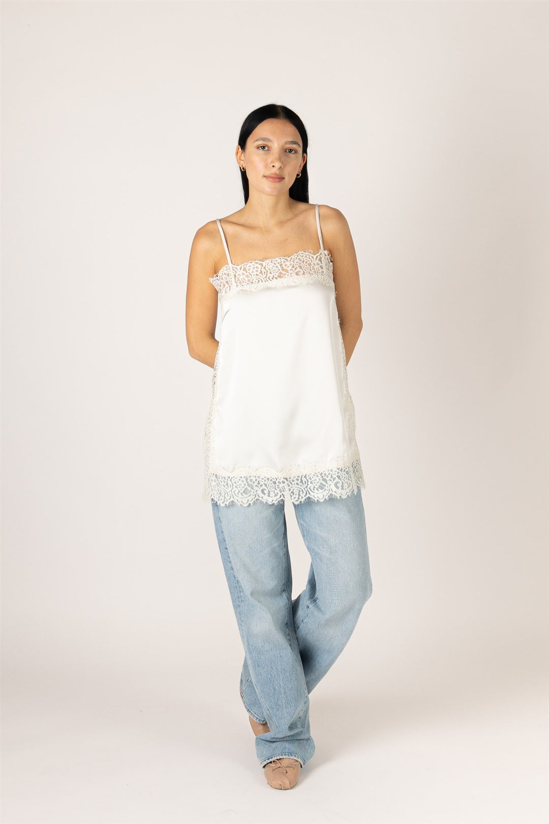 A woman modeling a white camisole top with delicate lace trim at the neckline and hem, paired with light-wash jeans, against a plain light beige studio background.