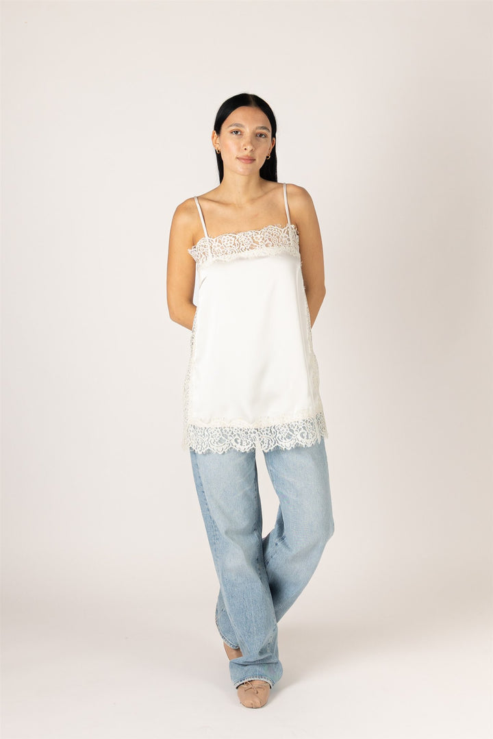 A woman modeling a white camisole top with delicate lace trim at the neckline and hem, paired with light-wash jeans, against a plain light beige studio background.