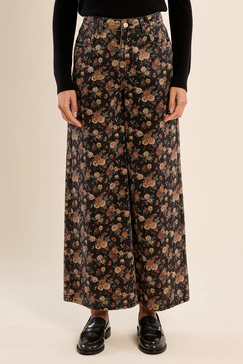 A model wearing high-waisted, wide-leg trousers with a black base and a dense floral print in shades of brown, beige, and muted orange. The model is also wearing a black long-sleeve top and black leather loafers, standing against a solid light beige background.