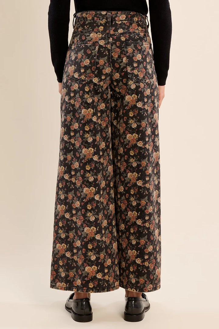 A person shown from the back wearing high-waisted, wide-leg pants with an all-over floral print on a black background, paired with a black long-sleeve top and black shoes, against a plain beige studio backdrop.