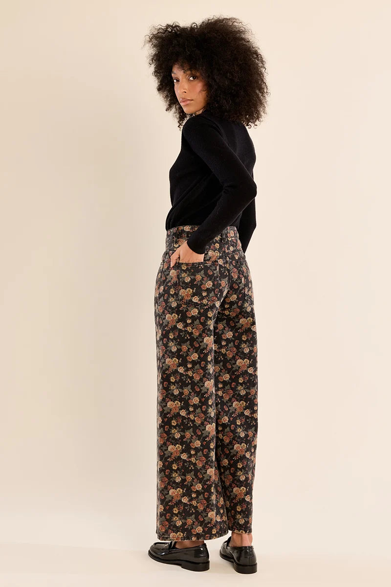 A woman stands in profile wearing a black long-sleeve top and wide-leg floral trousers with black loafers, photographed against a plain warm beige studio background.